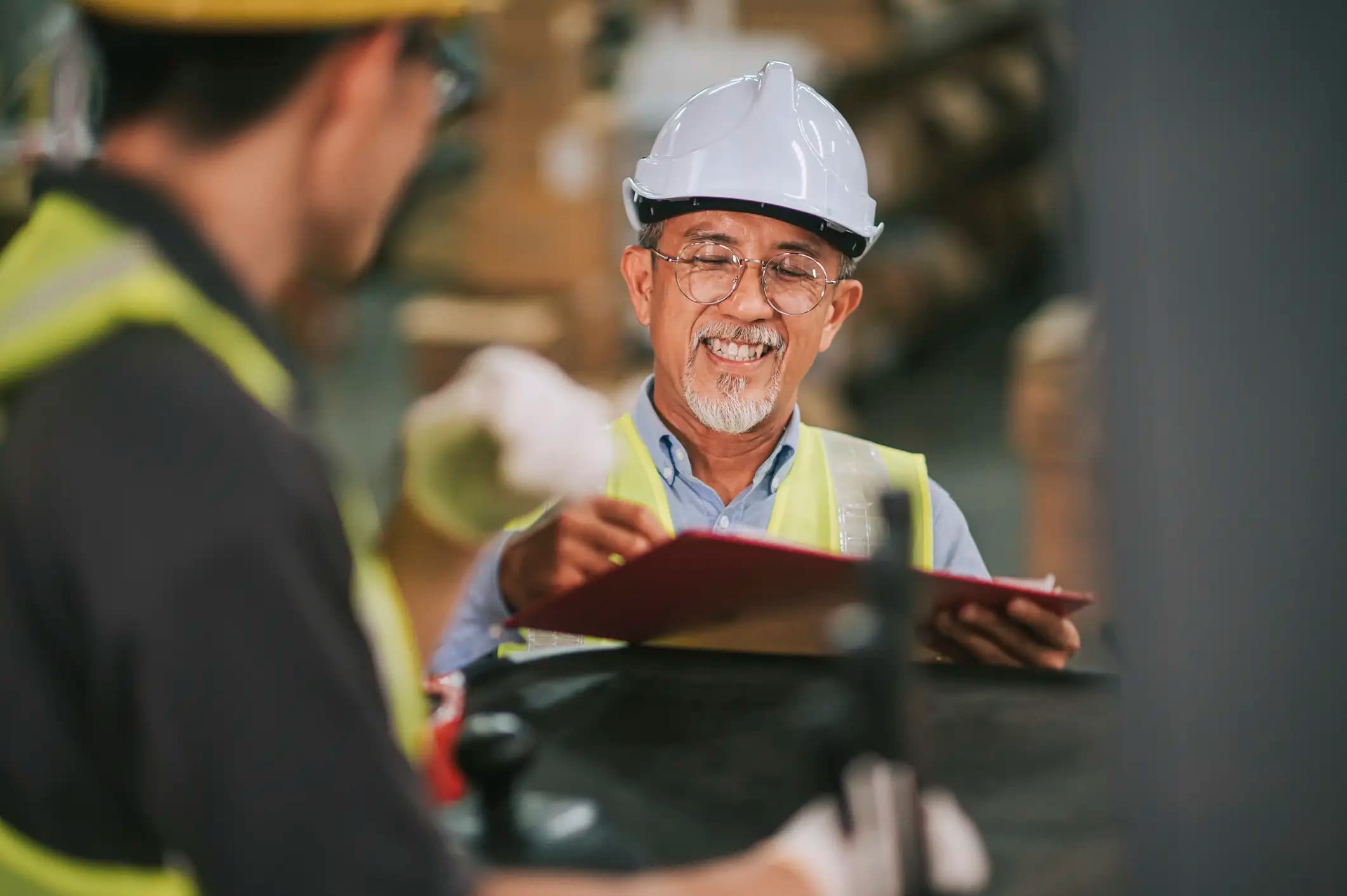 warehouse supervisor manager signing documents receiving goods from his coworker manual worker operating forklift working in industry factoryT wo mature men, in their 40-50s, working in a warehouse, wearing white hardhats and yellow safety vests. One of the workers is sitting on a forklift. The other is standing beside him holding a clipboard which they are both reading and discussing.